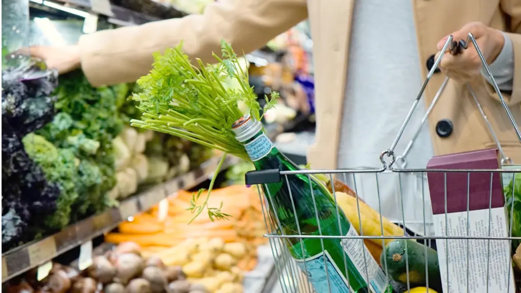 Eine Person hält einen Einkaufskorb mit verschiedenen gesunden Lebensmitteln in der einen Hand und greift mit der anderen Hand in ein Gemüseregal.