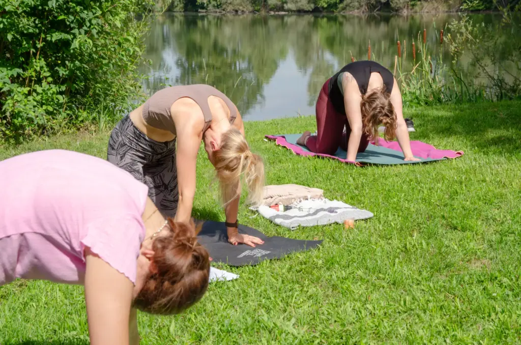 Bianca und zwei Teilnehmerinnen eines Retreats führen eine Yoga-Asana an einem Seeufer durch.